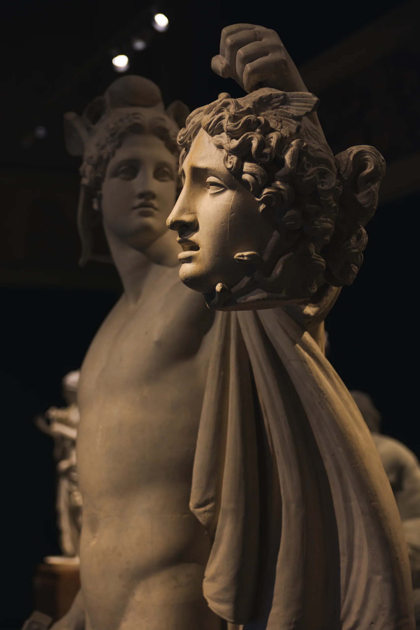 A marble group of a young man holding a severed head, another face visible behind his shoulder, lit warmly in a dark gallery.