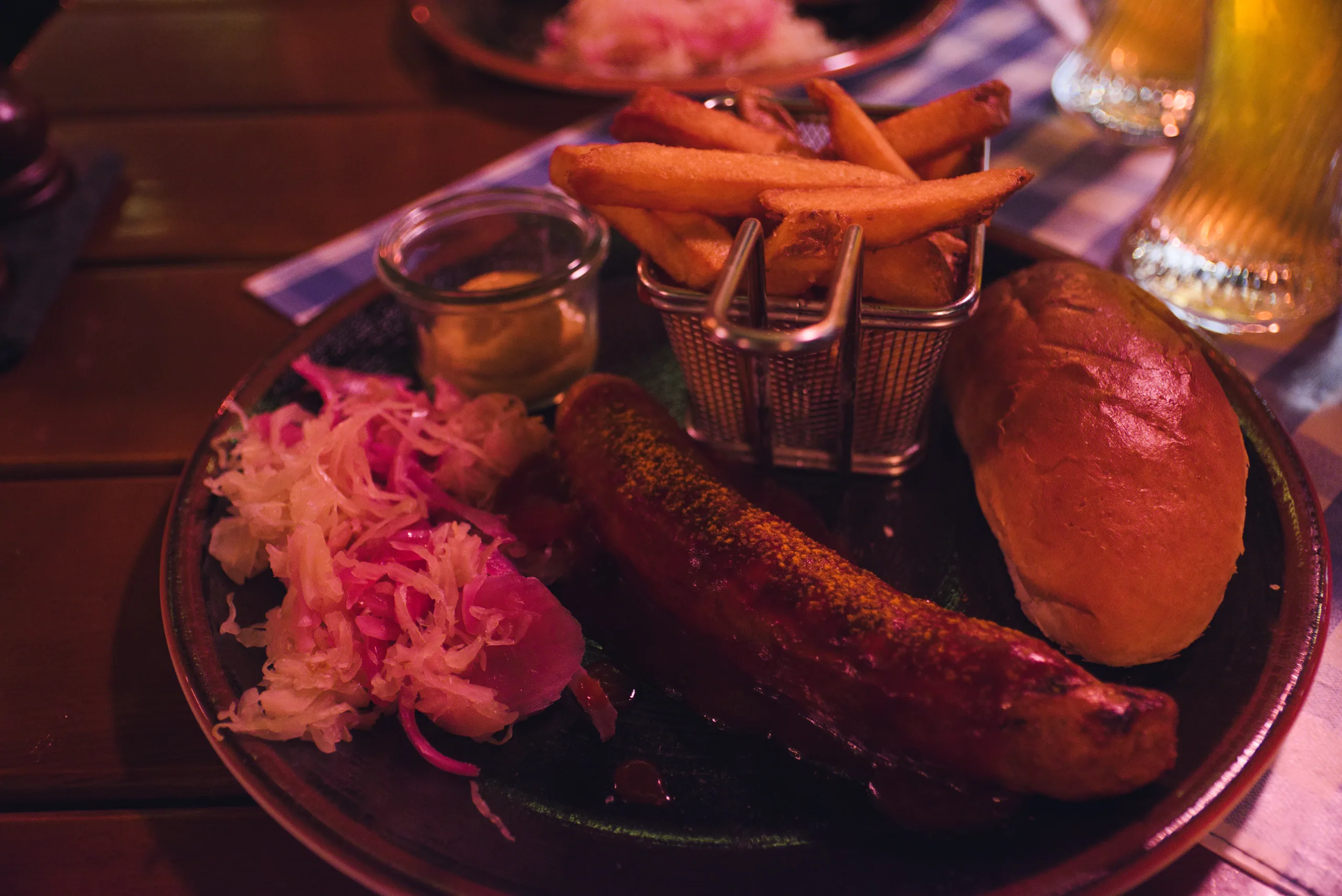 A pub plate of grilled sausage with pickled red cabbage, a bread roll and a basket of chips, a beer glass at the edge of the frame.