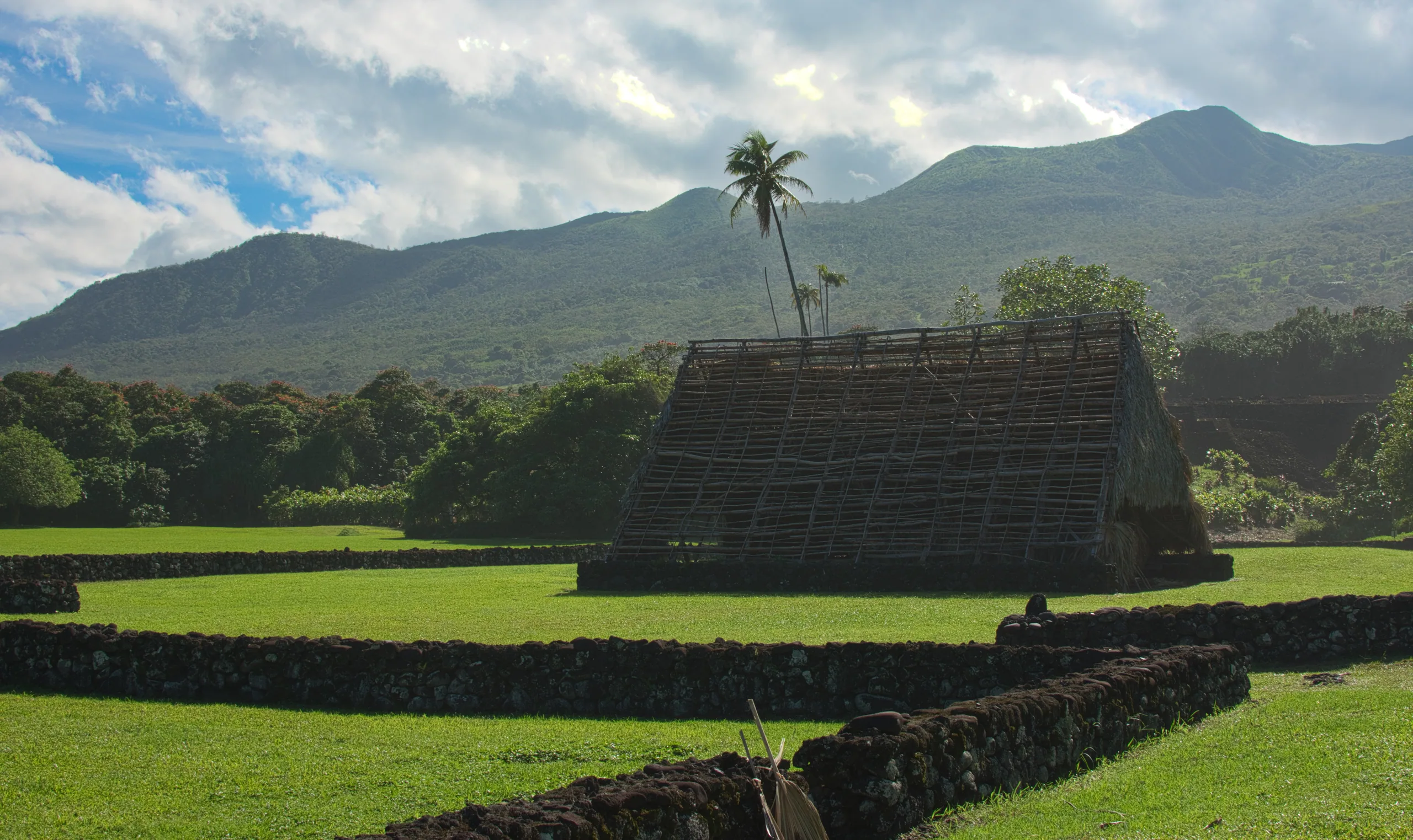 A thatched A-frame hale standing on a stone platform in a mown clearing, forested ridges rising behind.