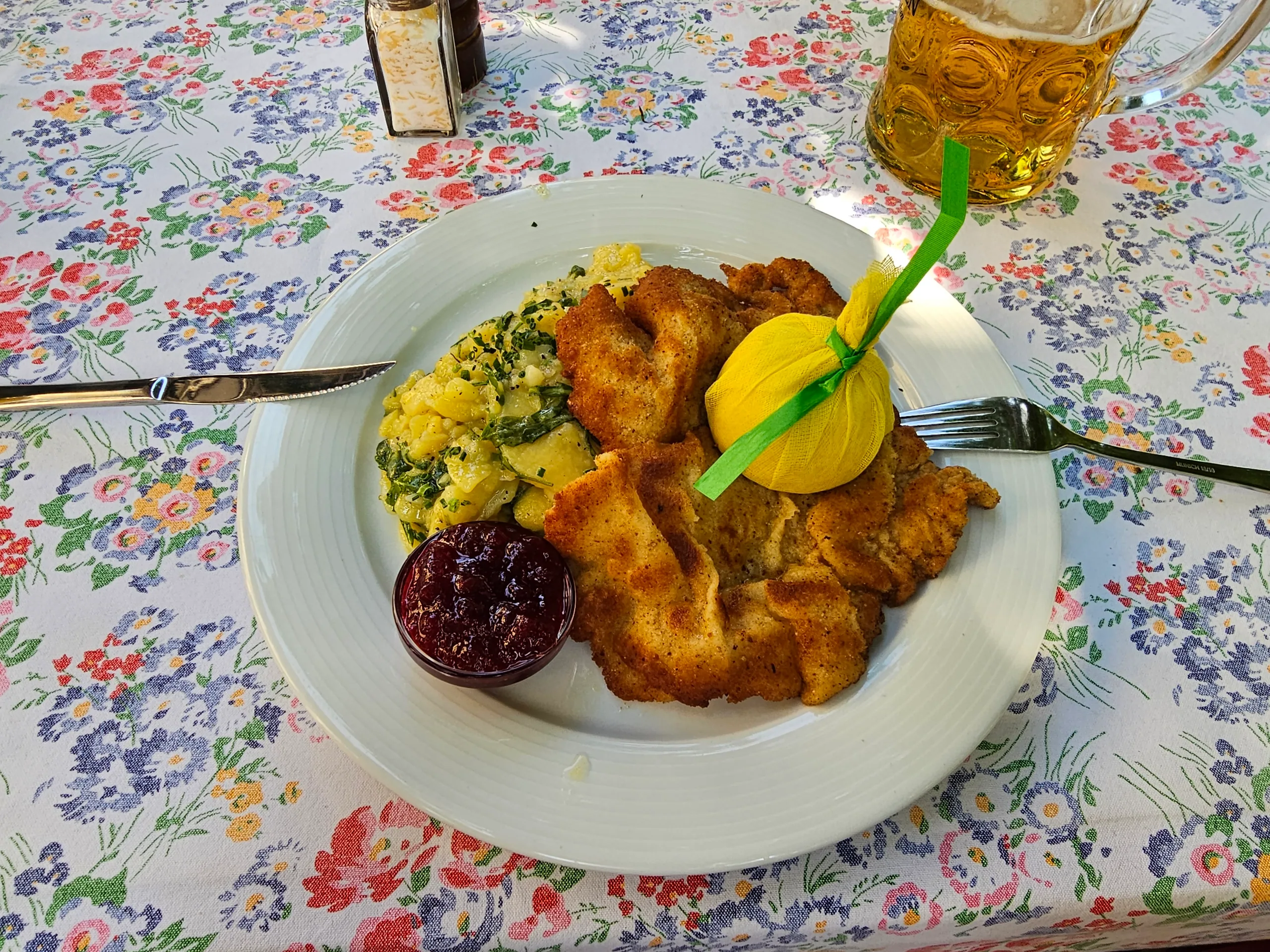 A breaded schnitzel with potato salad, a lemon wedge and lingonberry jam, on a floral tablecloth beside a glass of beer.
